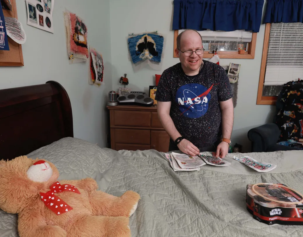 Person standing next to bed, in bedroom, holding artwork and smiling.